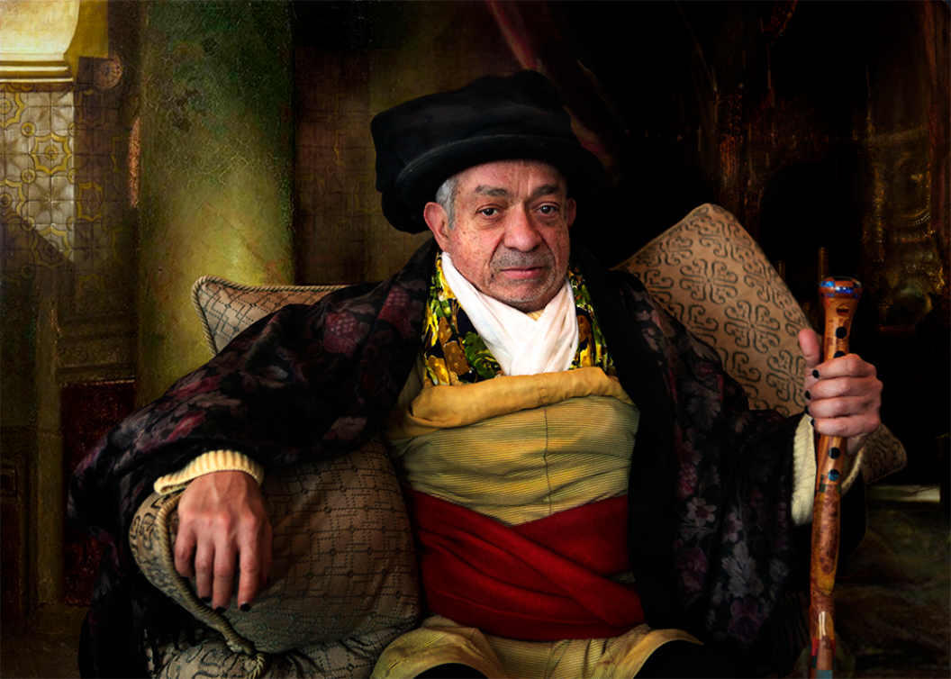 The author Rabih Alameddine costume of wrapped fabrics as he holds a decorated cane and sits against an abundance of pillows.
