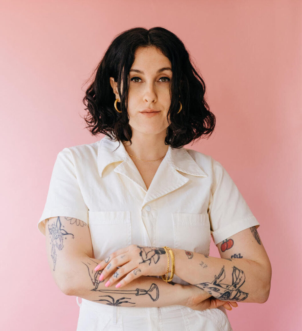 Pink background, light-skinned woman with chin-length wavy dark hair, tattooed arms folded, hoop earrings, white collared button up shirt. 