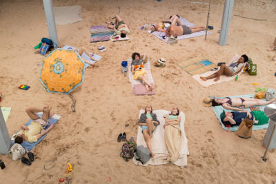 Areal view of a theatrical installation of a beach. People in beach attire lay on towels surrounded by miscellaneous beach gear. Above the beach scene is a scaffolding from which viewers can watch the performance.