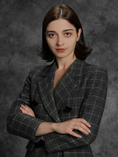 A young white woman with shoulder-length, brown hair and wearing a gray plaid blazer crosses her arms and looks directly into the camera in a waist-up portrait against a clouded gray photography studio backdrop.