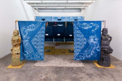 A tent made of indigo-dyed fabric panels, flanked on each side by gargoyle-like statues.