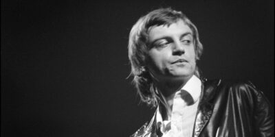 horiozontal, black-and-white headshot of The Fall's Mark E. Smith. He takes up the right half of the frame and looks off to the right with his lips pursed.