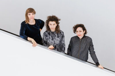 Three young women stand facing the camera on a staircase descending down to the right of the frame.