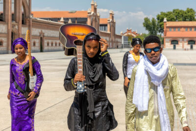 Three women (two carrying guitars) and one man walking through an empty square