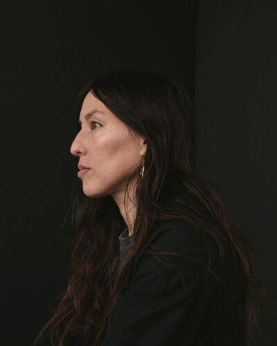 A young woman with long, dark hair sits in profile against a black backdrop.