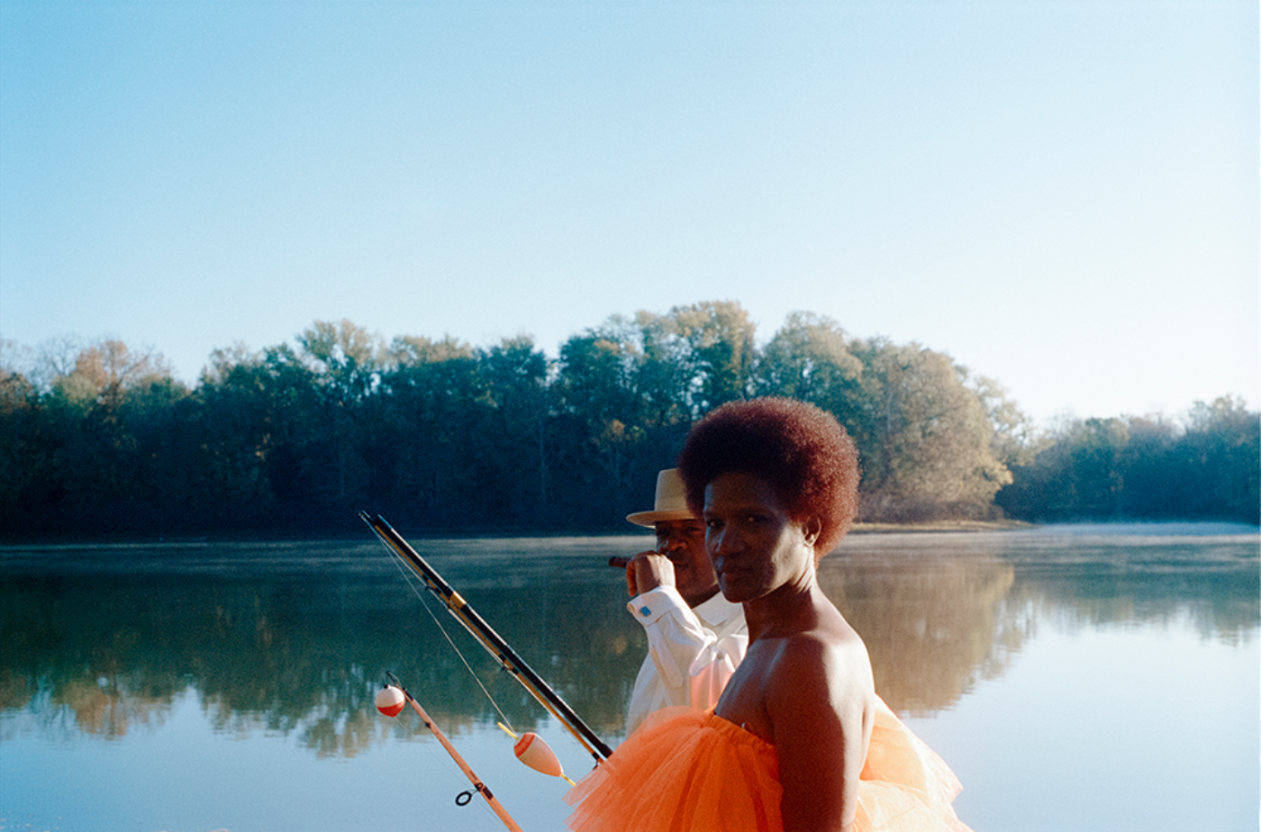 A Black woman in an orange dress and a Black man in a white suit and hat hold fishing rods with a lake in the background.