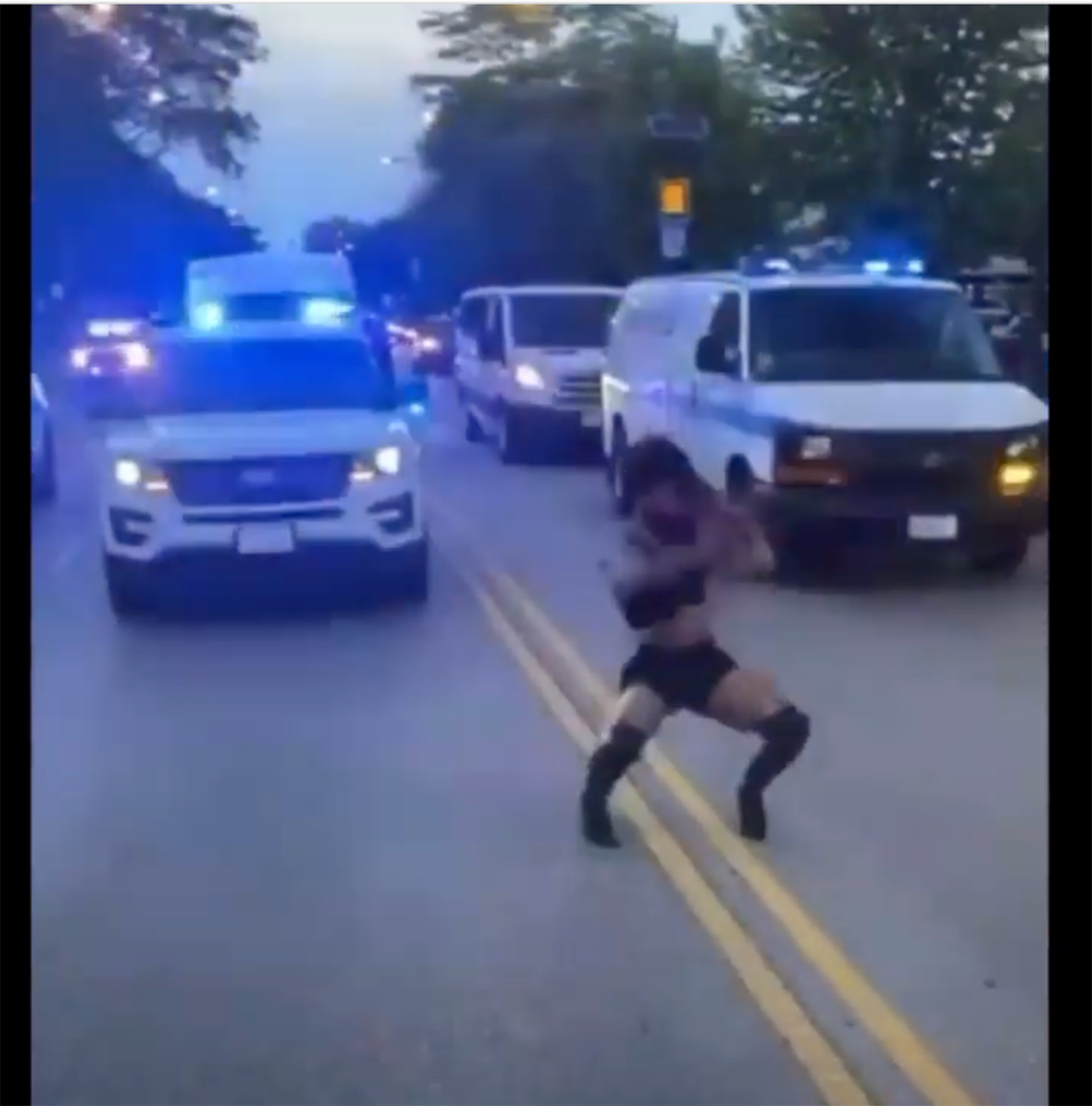 A Black woman in long black boots, shorts, and a tank top dances in the middle of the road in front of several police vehicles with their lights on.